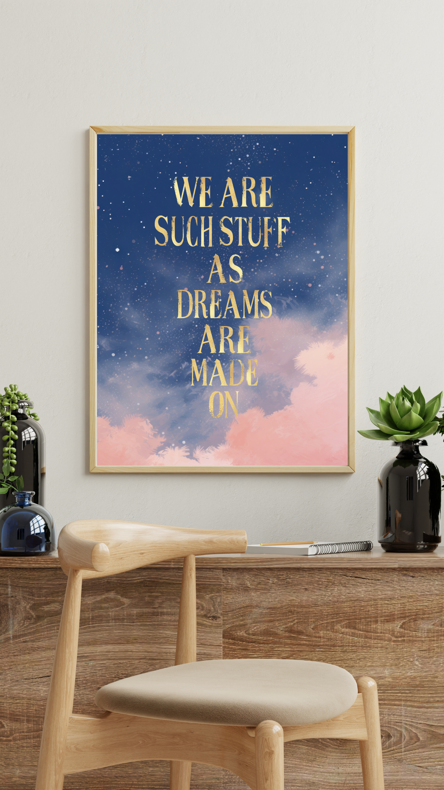 Framed wall art print with gold text reading “We are such stuff as dreams are made on” over a dreamy starry night sky with pink clouds, styled above a wooden console with plants and a modern chair.